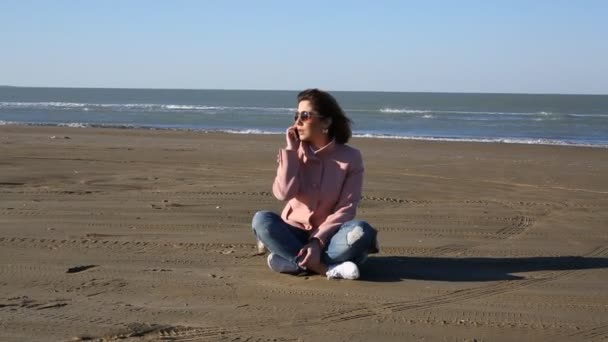 Милая девушка, сидящая в пиджаке на пляже и разговаривающая по телефону. Молодая женщина разговаривает по телефону на морском фоне, в Балтийском море. песчаная буря — стоковое видео