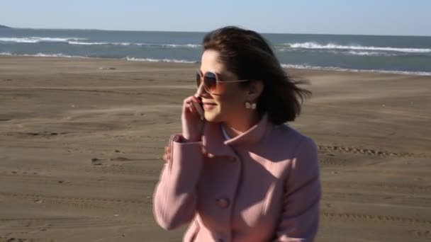Söt flicka sitter i jackan på stranden och prata i telefon. Ung kvinna prata i telefon på havet bakgrund, Östersjön. sandstorm — Stockvideo