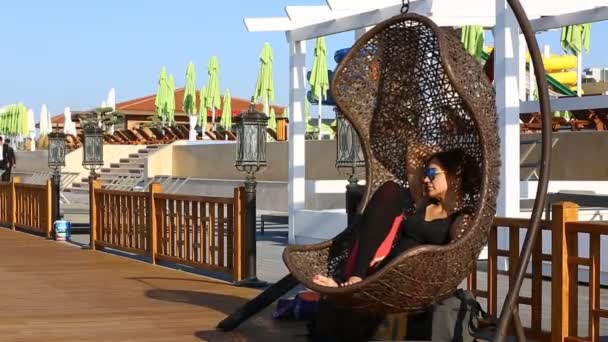 Femme assise sur un parc aquatique oscillant en osier. Femme à lunettes équitation dans une chaise en osier suspendu dans le parc ensoleillé journée d'été . — Video