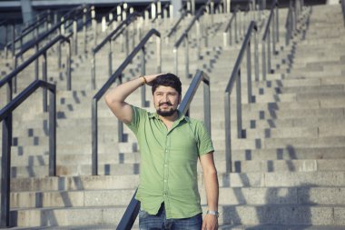 Yeşil tişörtlü ayakta adımlar üzerinde serin bıyıklı adam sakallı. Jest ciddi bakar.