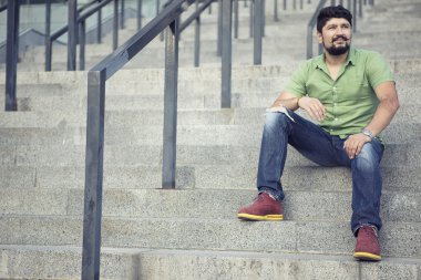 Yeşil tişörtlü ayakta adımlar üzerinde serin bıyıklı adam sakallı. Jest ciddi bakar.