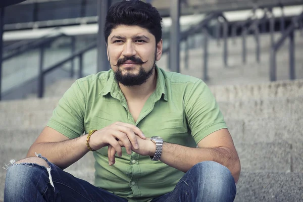 Yeşil tişörtlü ayakta adımlar üzerinde serin bıyıklı adam sakallı. Jest ciddi bakar.