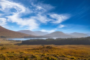 Yumuşak gün ışığıyla aydınlatılan geniş bir Highland vadisi. Huzurlu bir göl, ormanlı yamaçlar ve derin mavi gökyüzüne doğru süpürülen bulutlar. Manzara, vahşi doğa ve açık hava yaşam tarzı için mükemmel.