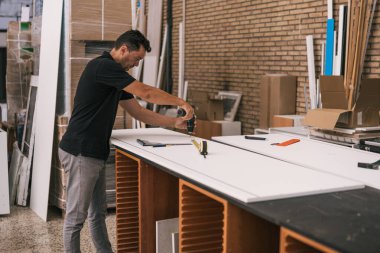 Atölyede beyaz bir yüzey üzerinde matkap kullanan bir adam.