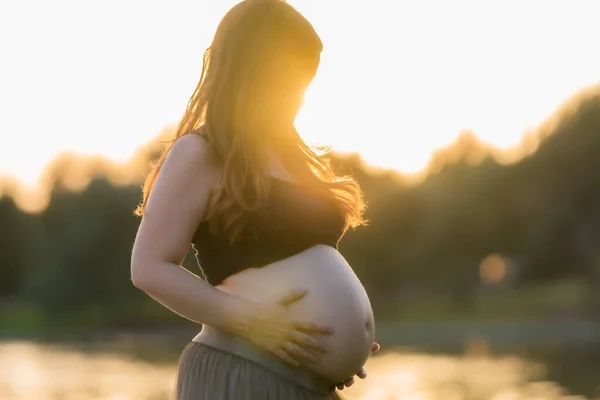 Strålar av solnedgång lyser upp en gravid kvinna när hon rör hennes mage utomhus — Stockfoto