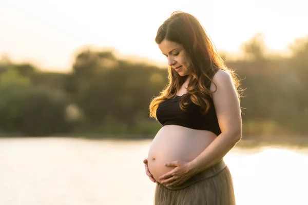 Gravid kvinna tittar och rör sin mage när hon står i en park — Stockfoto