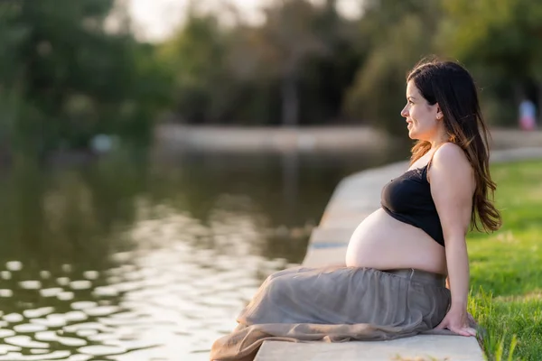 Gravid kvinna överväger floden i en park medan du sitter under solnedgången — Stockfoto