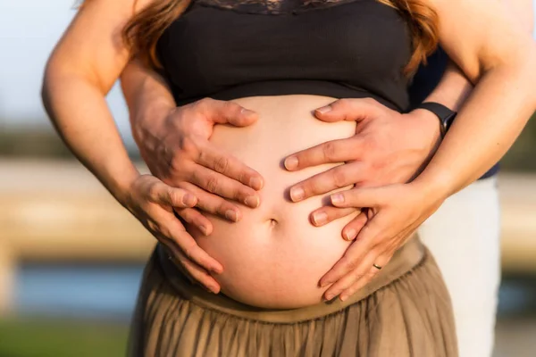 Närbild av fyra händer på magen på en gravid vuxen kvinna som står utomhus — Stockfoto