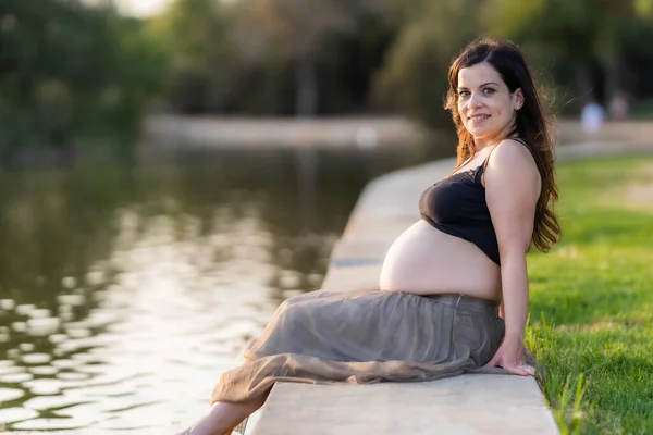 Mujer embarazada sonriente sentada al borde de un río en un parque durante el atardecer —  Fotos de Stock