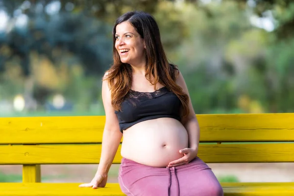 Leende gravid kvinna sitter på en parkbänk med en hand på magen i en solig dag — Stockfoto