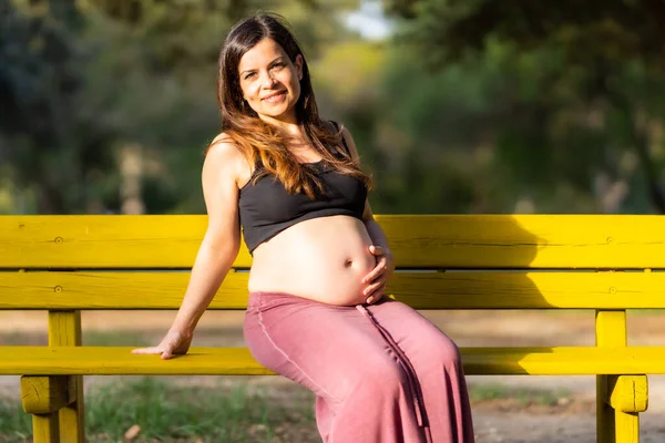 Mujer embarazada sentada en un banco del parque con una mano sobre su abdomen en un día soleado —  Fotos de Stock