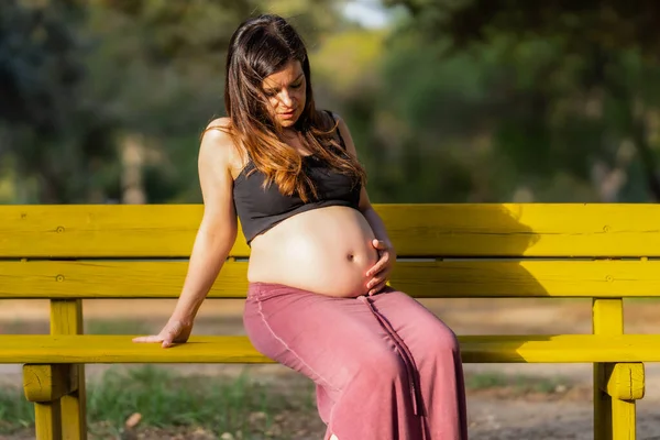 Gravid kvinna sitter på en bänk ser och röra sin mage i en solig dag — Stockfoto