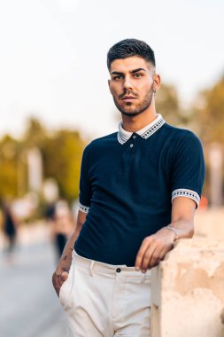 Modern yaz giysileri içinde duvara yaslanmış genç bir adamın dikey fotoğrafı.