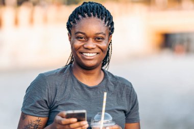 Kameraya gülümseyen Afro bir kadın. Elinde cep telefonu ve dışarıda bir titreme var.