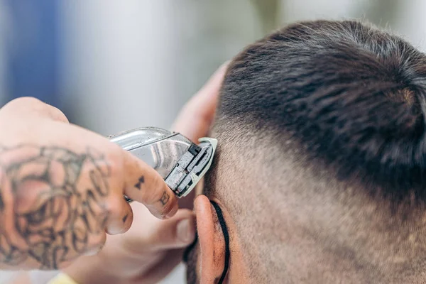 Mão tatuada usando uma máquina de barbear para cortar o cabelo de um cliente em uma barbearia — Fotografia de Stock