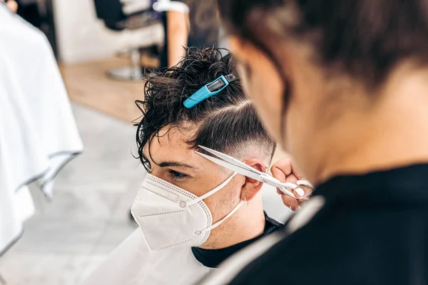 Jovem com máscara sentada enquanto uma mulher corta seu cabelo em um cabeleireiro — Fotografia de Stock