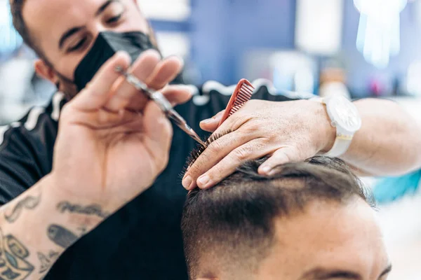 Barbeiro com máscara cortando o cabelo de um cliente usando tesoura em uma barbearia — Fotografia de Stock