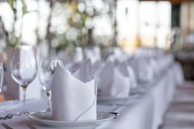 Modern bir restoranda misafirler için güzel bir masa hazırlanır. Beyaz peçeteler tabaklara güzelce katlanır ve bardaklar sıraya dizilir. Bu da yaklaşan bir olayı ya da özel bir yemeği işaret eder..