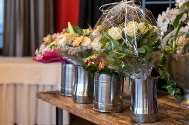 Açık plastiğe sarılı bir kaç baş döndürücü çiçek buketi pencere kenarındaki sevimli bir etkinlik sırasında ahşap bir masada parlak metalik vazolarda oturur..