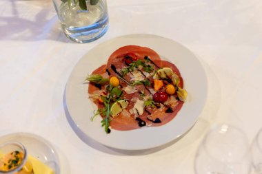 Güzel bir restoranda beyaz bir tabakta renkli bir salata sunulur. Tabakta dilimlenmiş et, yeşiller ve bir sos yağmuruyla süslenmiş süsler yer alıyor. Yemek masasında güzelce servis edilir..