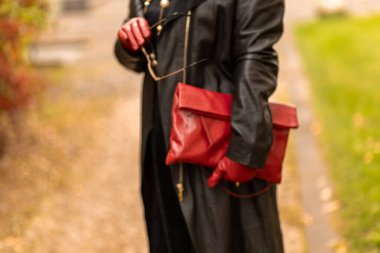 Uzun siyah bir ceket, kırmızı eldivenler giymiş ve elinde kırmızı bir çantayla sonbahar yapraklarıyla dolu bir parkta gezen bir kadın. Atmosfer sakin, sonbahar renklerini sergiliyor..