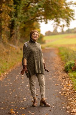 Yaşlı bir kadın sonbahar yapraklarıyla kaplı manzaralı bir yolda mutlu mesut duruyor. Sıcak, şık bir kıyafet giyiyor ve doğada huzurlu bir anın tadını çıkarıyor..