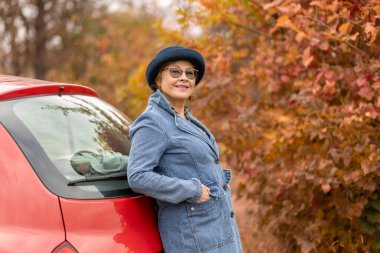 Kırmızı bir arabaya yaslanırken kot ceket giyen ve şık bir şapka takan bir kadın gülümsüyor. Canlı sonbahar yaprakları, açık bir günde sıcaklık ve renk saçan güzel bir zemin oluşturur..