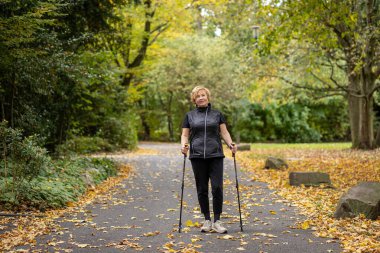 Bir kadın, rengarenk sonbahar yapraklarıyla çevrili huzurlu bir park yolunda güvenle yürür. Destek için yürüyüş direklerini kullanıyor, sakin bir ortamda doğanın güzelliğinin tadını çıkarıyor..
