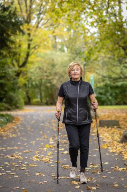 Son sınıf öğrencisi bir kadın güvenli bir şekilde parkta yürür, baston kullanır. Sonbahar yaprakları zemini kaplayarak, sarı ve turuncu renklerle dolu canlı bir ortam yaratır..