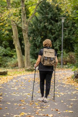 Bir insan, sonbahar parkında sırt çantası takarken baston kullanarak huzurlu bir yolda yürür. Renkli yapraklar uzun ağaçların altındaki zemini kaplıyor..