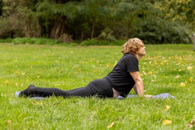 Kısa saçlı yaşlı bir kadın yemyeşil bir çayırda yoga yapıyor. Etrafı ağaçlar ve renkli sonbahar yapraklarıyla çevriliyken minder üzerinde poz veriyor..