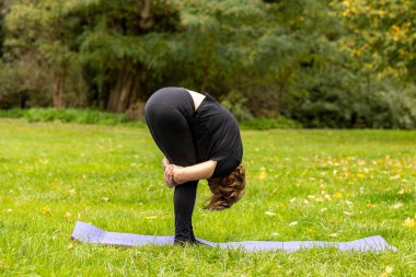 Yaşlı bir kadın huzur dolu bir parkta nazikçe yoga yapıyor. Etrafı yeşillik ve ağaçlarla çevrili, sakin bir duruşla öne eğiliyor, doğal huzurun tadını çıkarıyor..