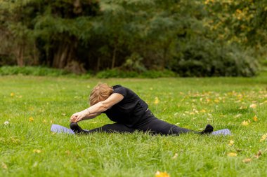 Yaşlı bir kadın ağaçlarla çevrili sakin bir çimenlikte yoga yaparken derin bir esneme hareketi yapıyor. Sahne sonbaharda doğanın güzelliğini yakalar..
