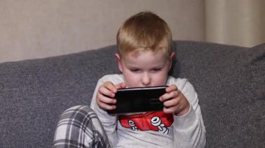 Bir çocuk koltuğa oturur ve mobil bir cihaza odaklanır. Çocuk bir oyunla meşgul, konsantrasyon ve ilgi gösteriyor. Oturma odasında olur böyle şeyler..