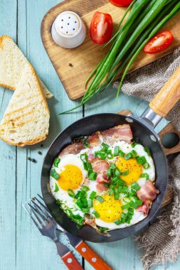 Çeşnili yumurta, yeşil soğan ve domuz pastırması. Kırsal ahşap arka planda dökme demir tavada. Üst görünüm düzlüğü.