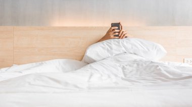 Man waking up and use smartphone or mobile phone on the bed in white blanket at morning time, social lifestyle concept