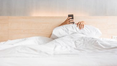 Man waking up and use smartphone or mobile phone on the bed in white blanket at morning time, social lifestyle concept