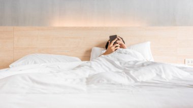 Man waking up and use smartphone or mobile phone on the bed in white blanket at morning time, social lifestyle concept