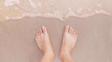 Tatilde deniz dalgalarıyla kumsalda duran ayaklar rahatlatıcı ve seyahat konsepti.