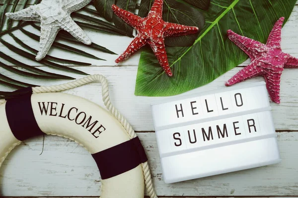 Hello Summer text in light box with green leave, starfish and lifebuoy decoration on wooden background