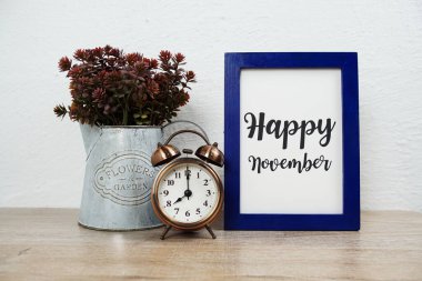 Alarm saati ve tahta masa ve beyaz duvar zemininde yapay bitki süslemesi bulunan mutlu Kasım tipografi metinleri