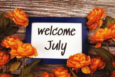 Ahşap zemin üzerinde çiçek süslemesi olan mavi kenar çerçeveli Temmuz ayına hoş geldiniz metni