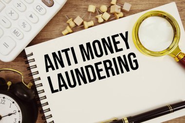 Not defterine kalem, alarm saati, büyüteç ve ahşap zemin üzerinde hesap makinesi ile yazılmış Para Aklama Karşıtı metin