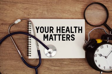 Not defterinize steteskop, büyüteç ve ahşap arka planda çalar saat ile yazılmış Sağlık Sorunları metniniz