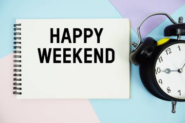 Çalar saatin düz olduğu not defterine yazılmış Mutlu Hafta Sonu metni çok renkli pastel arka planda yatıyordu
