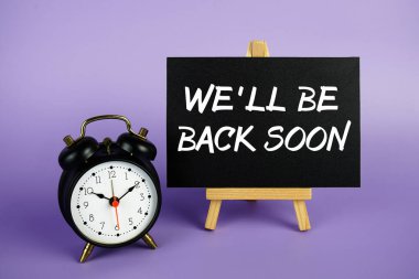 Birazdan döneriz. Mor zemin üzerinde resim sehpası ve çalar saatle.