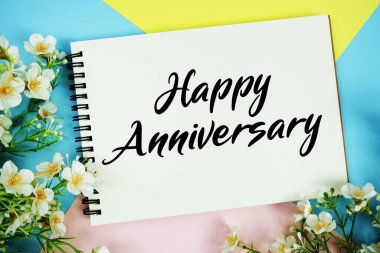 Çiçek dekorasyonlu not defterine yazılan mutlu yıldönümü metni çok renkli pastel arka planda yatıyordu