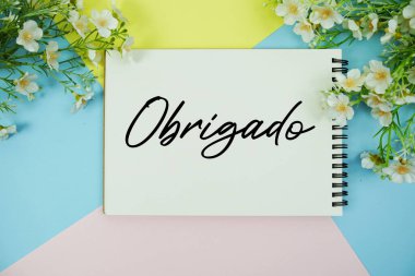Çiçek dekorasyonlarıyla birlikte not defterine yazılmış Obrigado metni çok renkli pastel arka planda yatıyordu