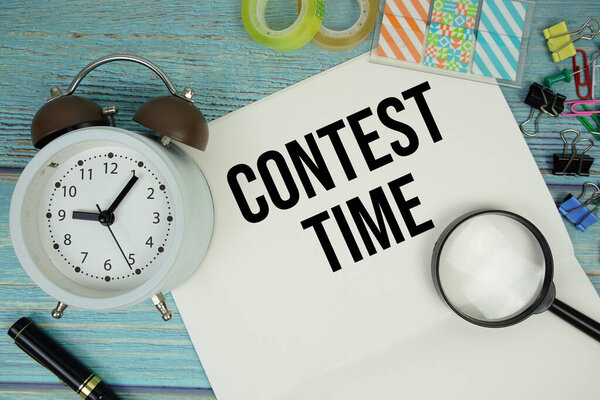 Contest Time text on a paper surrounded by office supplies, a magnifying glass, and a clock on a blue wooden surface