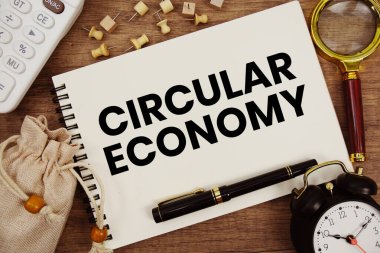 Kalem, alarm saati, büyüteç ve ahşap zemin üzerinde hesap makinesi ile not defterine yazılmış dairesel ekonomi metni